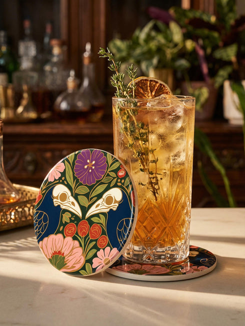 A square drink coaster with a high-gloss finish and natural cork backing. The centralized, symmetrical design features two detailed, cream-colored avian bird skulls facing inward. They are framed by lush botanical flora, including a large purple flower, blush pink blooms, rust-red roses, and green ferns, all set against a deep navy blue background with gold linework accents. Functional gothic, whimsigoth, and dark academia drinkware designed by the Grimoire and Goblet brand.