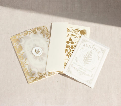 a luxury Grimoire and Goblet ritual card set. A 5x7 cream-colored folded card features an intricate, art-nouveau-style gold foil illustration of a Juniper plant, with the words "Juniper: Cleansing & Fortune." The card is presented with a translucent vellum envelope, which has a gold floral border and a white adhesive wax seal. Inside, the card reveals a vellum-wrapped keepsake, which unfurls to a 4x6 ritual card printed on thick, 160 GSM handmade cotton paper with a rough, deckled edge.