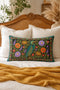 Heron lumbar pillow cover featuring a teal wading bird with gold detailing standing among orange marigolds, golden chrysanthemums, purple peonies, lavender sprigs, and coral red blooms with green foliage on a deep green black panel. A teal border with gold rope trim and a folk style band of red rosettes frames the design. Rectangular 20 x 12 inch cushion case with hidden zipper, the same tapestry look design printed on both sides in smooth, linen feel polyester fabric.