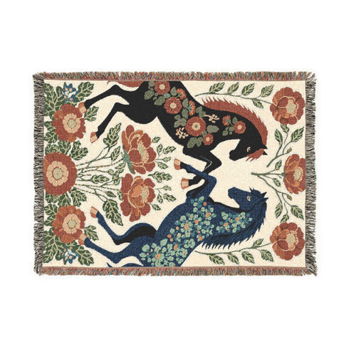 A woven cotton throw blanket by Grimoire and Goblet featuring two rearing horses facing each other in a symmetrical folk art design. One horse is charcoal black with rust floral detailing; the other is navy blue with teal floral detailing. They are framed by large rust-colored peonies and green leafy vines on a cream background. The blanket features a multicolor fringe edge. Perfect for dark academia and cottagecore aesthetics.