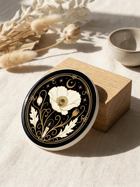 A set of four round 4-inch ceramic coasters featuring a glossy 2D flat-printed Art Deco botanical design. Each coaster displays a different cream-colored flower including a moonflower, foxglove, poppy, and iris, set against a solid black background. The floral motifs are framed by intricate gold linework, crescent moons, four-pointed stars, and mystical symbols like a candle and an all-seeing eye. The coasters have a smooth white edge and are finished with a natural brown cork backing.
