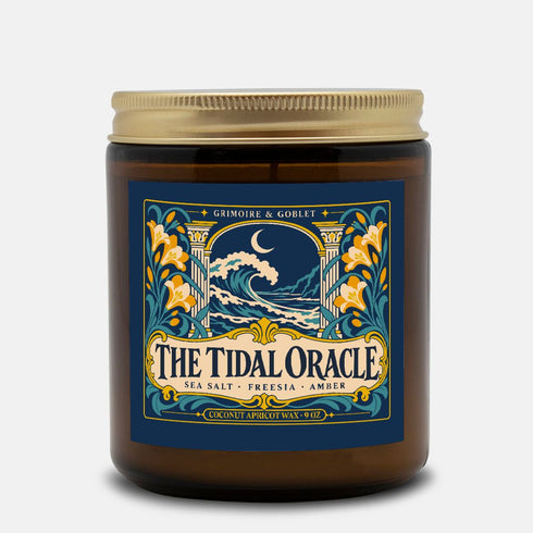 A 9 oz amber glass jar candle with a brushed gold screw lid and a wraparound deep navy label. The label shows a cresting wave beneath a crescent moon, framed by two classical columns and an arched window, with golden art nouveau freesias climbing both sides on teal stems, all inside a fine gold border. A cream banner reads The Tidal Oracle, with Grimoire & Goblet above and Sea Salt, Freesia, Amber and Coconut Apricot Wax, 9 oz below. A celestial coastal design.