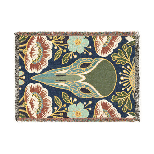 A midnight navy woven cotton blanket featuring a large central raven skull surrounded by rust and ochre flowers, and sage green ferns. Crescent moons are located in the four corners. The blanket has a colorful fringed edge and a textured, loomed appearance. Displayed as a premium gothic botanical home decor piece by Grimoire and Goblet.