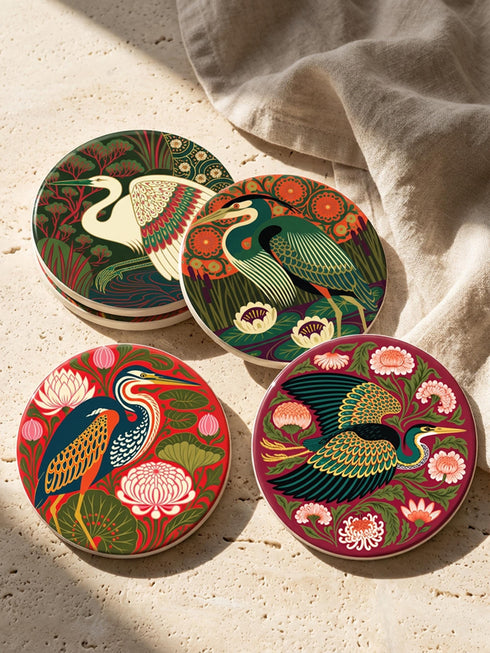 This set of four round ceramic coasters features a glossy, smooth, high-fidelity flat-printed surface. Each piece displays highly detailed Art Nouveau style illustrations of herons and egrets surrounded by stylized water lilies, chrysanthemums, and rich foliage. The vibrant, intricate color palette includes deep emerald greens, rich crimson reds, navy blues, and golden yellows. The sides are smooth, unprinted white ceramic, and the underside is fitted with a natural brown cork backing.