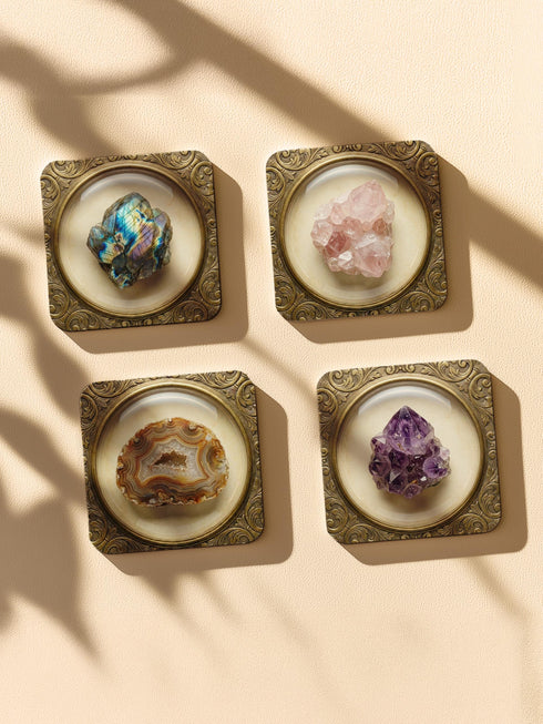Grimoire and Goblet set of four square glossy coasters featuring high-resolution printed illustrations of raw crystals: purple amethyst, pink rose quartz, iridescent labradorite, and banded agate. Each gemstone is depicted inside a printed faux antique brass baroque frame with intricate scrollwork, set against a cream parchment background. The design is a flat 2D trompe l'œil print on hardboard, creating a realistic 3D specimen box illusion.
