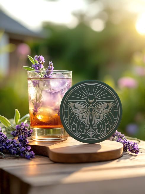 A round, dark hunter green ceramic drink coaster with a non-slip cork back. The top surface features an intricate, engraved-style celestial moth design in a light beige or off-white color. The luna moth is centered under a crescent moon and a radiant sunburst. The design is symmetrical and includes stars and geometric line art, evoking a gothic, witchy, and spiritual aesthetic. The coaster has a matte finish and is designed as a piece of functional art.