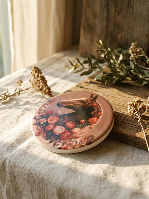 A round glazed ceramic coaster by Grimoire and Goblet featuring a vintage Victorian horse portrait design. The coaster has a high gloss porcelain-like finish and a natural cork bottom for furniture protection. The artwork depicts a dark bay horse head in profile surrounded by moody botanical flowers in shades of rust and deep red, evoking a dark academia and gothic cottagecore aesthetic. This 4x4 inch tile serves as a protective drink mat or decorative equine art accent.