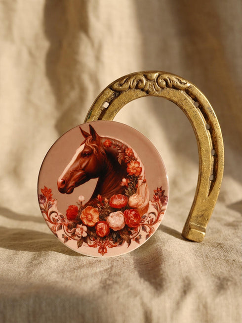 A round glazed ceramic coaster by Grimoire and Goblet featuring a vintage Victorian horse portrait design. The coaster has a high gloss porcelain-like finish and a natural cork bottom for furniture protection. The artwork depicts a dark bay horse head in profile surrounded by moody botanical flowers in shades of rust and deep red, evoking a dark academia and gothic cottagecore aesthetic. This 4x4 inch tile serves as a protective drink mat or decorative equine art accent.