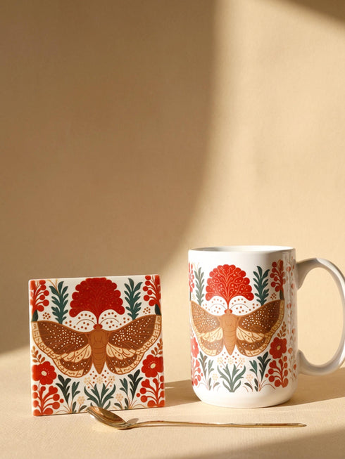 A detailed close-up of a luxury ceramic mug and matching square coaster set from Grimoire and Goblet. The items feature a vintage-style scientific illustration of a large moth with intricate wing patterns in earth tones of rust red and moss green, resting on stylized botanical foliage. The aesthetic is Dark Academia and Victorian Gothic. The mug is a large 15oz white ceramic vessel with a sturdy C-handle. The coaster is a glazed ceramic tile with a glossy finish.