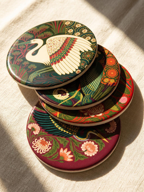 This set of four round ceramic coasters features a glossy, smooth, high-fidelity flat-printed surface. Each piece displays highly detailed Art Nouveau style illustrations of herons and egrets surrounded by stylized water lilies, chrysanthemums, and rich foliage. The vibrant, intricate color palette includes deep emerald greens, rich crimson reds, navy blues, and golden yellows. The sides are smooth, unprinted white ceramic, and the underside is fitted with a natural brown cork backing.