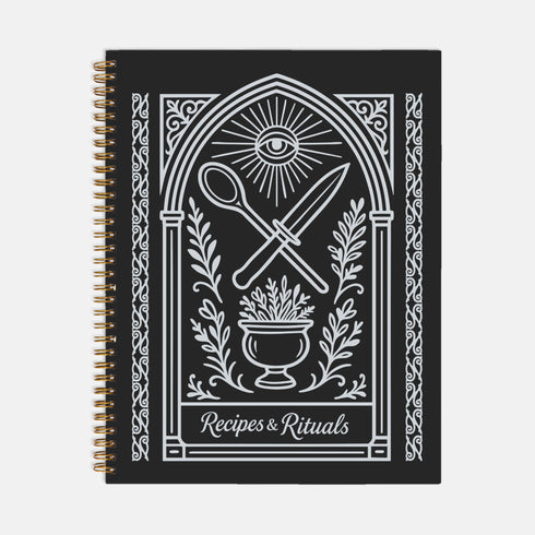 A black spiral-bound hardcover recipe journal lies flat. The cover is matte black with a white line-art design titled "Recipes & Rituals." The art nouveau style illustration features an all-seeing eye under a sunburst, framed by an ornate arch. Below the eye, a crossed spoon and knife are centered above a mortar and pestle filled with herbs. The entire design is bordered by intricate botanical flourishes. The notebook is 8.5 x 11 inches with a wire-o binding, a lay-flat cookbook for kitchen use.