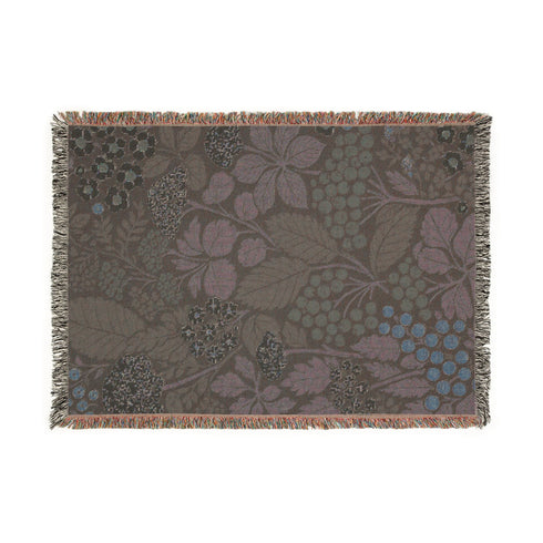 A Grimoire and Goblet luxury woven throw blanket featuring a dark botanical design. The Jacquard cotton tapestry showcases cream elderflower clusters and rust-colored berries against a deep black background. Sage green leaves interweave throughout the floral pattern. The blanket is finished with multicolored fringe edges. A moody cottagecore aesthetic piece ideal for dark academia decor.