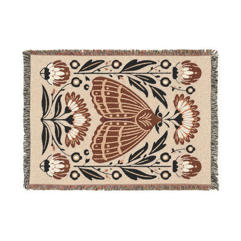 Grimoire and Goblet woven jacquard cotton blanket / wall tapestry featuring a centered folk-art moth framed by symmetrical wildflowers and leafy stems. Natural oatmeal-beige base with rust/terracotta and cocoa-brown moth detail, charcoal-black botanicals, and warm ivory highlights. Finished with a 2-inch multicolor fringe and a reversible color-negative back. Available in 52x37, 50x60, and 60x80 inches as a throw, bed cover, or statement wall hanging.