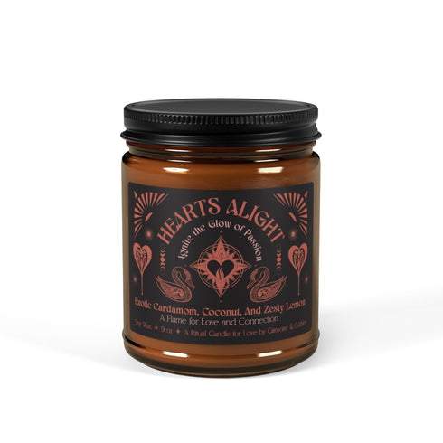 A 9 oz amber glass candle featuring a black label with rose gold art depicting hearts and swans. The label reads "Hearts Alight" and lists cardamom, coconut, and lemon fragrance notes. A cotton wick burns at the center.