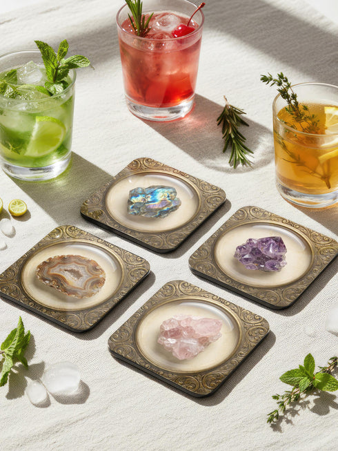 Grimoire and Goblet set of four square glossy coasters featuring high-resolution printed illustrations of raw crystals: purple amethyst, pink rose quartz, iridescent labradorite, and banded agate. Each gemstone is depicted inside a printed faux antique brass baroque frame with intricate scrollwork, set against a cream parchment background. The design is a flat 2D trompe l'œil print on hardboard, creating a realistic 3D specimen box illusion.
