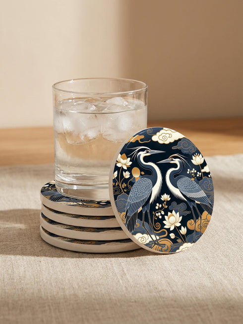 A round 4-inch drink coaster by Grimoire and Goblet. The coaster features a highly reflective glossy top surface displaying two Great Blue Herons, lotus flowers, and stylized clouds in metallic antique gold and slate blue against a solid, deep navy blue background. The edges reveal a natural brown, porous cork backing underneath. The intricate design resembles vintage Chinoiserie and botanical natural history art, perfectly suited for protecting tables from beverages.