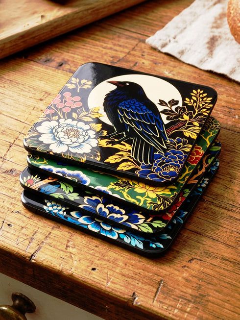 Grimoire and Goblet set of 4 glossy square coasters featuring Chinoiserie style raven illustrations. 1: Navy blue with blue floral pattern and raven. 2: Navy background with black raven, white full moon, and red berries. 3: Moss green background with blue bird and yellow chrysanthemums. 4: Black background with teal-winged raven and red peonies. MDF hardboard with cork backing. Flat vector botanical art style.