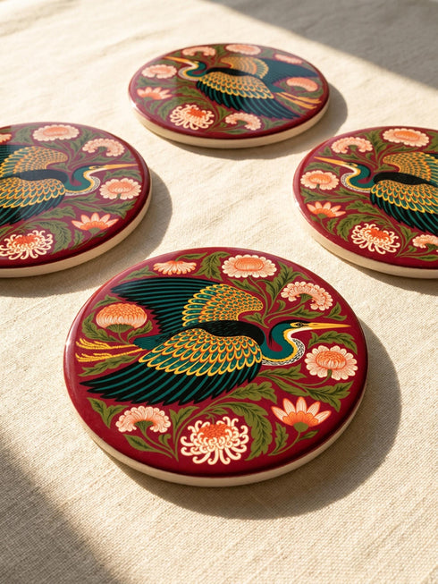 A round, 4-inch ceramic drink coaster featuring a glossy, flat-printed Art Nouveau design. The central motif is a flying heron with teal plumage and scalloped golden wing feathers, surrounded by stylized winding olive green foliage, peach chrysanthemums, and botanical blooms. The artwork is set against a solid, deep blood-red background. The coaster has a smooth white edge and a natural brown cork backing. Available as a single coaster or in matching sets of four or six.