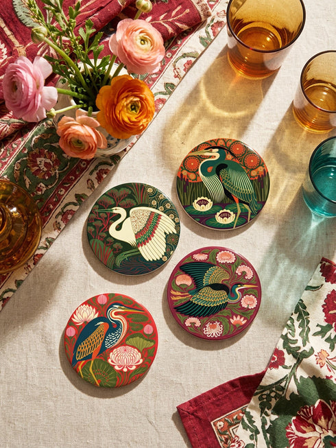 This set of four round ceramic coasters features a glossy, smooth, high-fidelity flat-printed surface. Each piece displays highly detailed Art Nouveau style illustrations of herons and egrets surrounded by stylized water lilies, chrysanthemums, and rich foliage. The vibrant, intricate color palette includes deep emerald greens, rich crimson reds, navy blues, and golden yellows. The sides are smooth, unprinted white ceramic, and the underside is fitted with a natural brown cork backing.