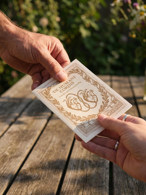 A vintage-style greeting card titled "DE HUMANI CORDIS" featuring a medical illustration of two anatomical hearts connected by a single looped blood vessel. The hearts are drawn in a sepia and crimson woodcut style on a cream background mimicking aged parchment. Text below reads "Fig. 14: The Twin Pulse." The card evokes a dark academia or cottagecore aesthetic. Inside text reads "Two different beats, one shared rhythm.
