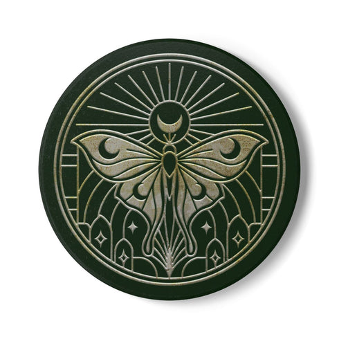 A round, dark hunter green ceramic drink coaster with a non-slip cork back. The top surface features an intricate, engraved-style celestial moth design in a light beige or off-white color. The luna moth is centered under a crescent moon and a radiant sunburst. The design is symmetrical and includes stars and geometric line art, evoking a gothic, witchy, and spiritual aesthetic. The coaster has a matte finish and is designed as a piece of functional art.