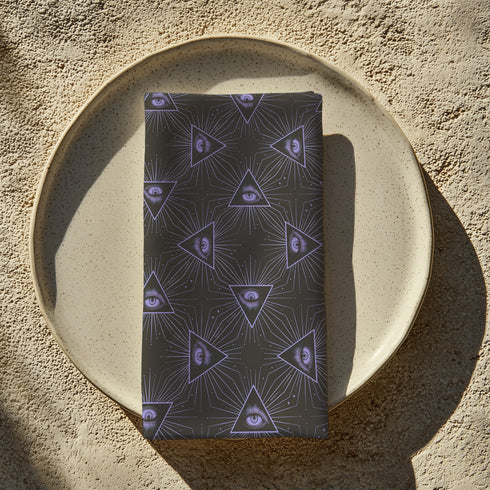Set of four charcoal-colored cloth napkins with a repeating pattern of triangular all-seeing eyes in purple, surrounded by radiating lines and small star-like accents.
