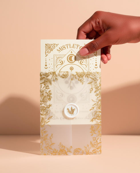 A Grimoire & Goblet luxury Mistletoe ritual card set. The 5x7 cream greeting card features gold celestial designs framing “Mistletoe” and “Love, Protection & New Beginnings.” Shown with a vellum wrap printed in gold florals, a white wax seal with mistletoe, and a premium envelope. Includes a 4x6 ritual insert on handmade deckled paper titled “Ritual for Peace and Protection.” A spiritual Yule stationery set designed as a luxurious unboxing experience.