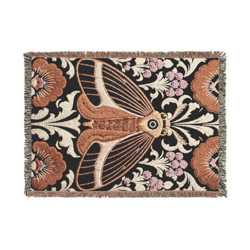 Woven cotton throw blanket with fringed edges and a black ground. At the center is a large moth with copper, brown, and cream wings marked by fine stripes and a concentric eyespot, with feathered antennae. Cream acanthus scrollwork fans out around it in the Art Nouveau manner, joined by clusters of lilac and mauve flowers and copper-brown blossoms with pale rays at the top and bottom. The pattern is jacquard woven, and the reverse shows a mirrored, softer version of the design.