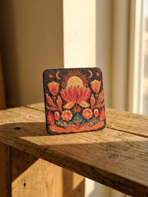 A Grimoire and Goblet drink coaster featuring a rustic faux burlap texture print. The coaster displays a gothic botanical design in dark charcoal ink on a tan jute-like polyester background. The smooth fabric surface mimics a woven look. Ideal for Dark Academia decor and protecting tables from spills. The design includes a central motif with vintage styling.