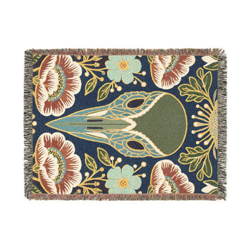 A midnight navy woven cotton blanket featuring a large central raven skull surrounded by rust and ochre flowers, and sage green ferns. Crescent moons are located in the four corners. The blanket has a colorful fringed edge and a textured, loomed appearance. Displayed as a premium gothic botanical home decor piece by Grimoire and Goblet.