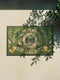 A rectangular doormat featuring a printed faux embroidery design on a muted olive green background. The symmetrical artwork centers on an all-seeing eye inside a circular medallion with a radiating sunburst pattern. An antique gold banner curves above reading WHAT YOU BRING TO THIS DOOR. A matching banner below reads FOLLOWS YOU HOME. The central eye is flanked by detailed botanical fern fronds and delicate wildflowers. The mat is finished with a sleek black fabric border trim.