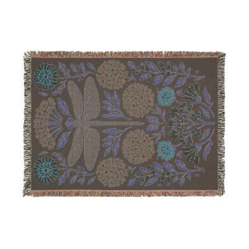 A Grimoire and Goblet Jacquard woven cotton blanket featuring a large central sage green dragonfly. The insect is surrounded by symmetrical clusters of slate blue hydrangeas and muted coral zinnia flowers against a black background. The blanket has a tactile woven texture and is finished with multicolor fringe edges. The design reflects a dark academia and vintage botanical aesthetic.