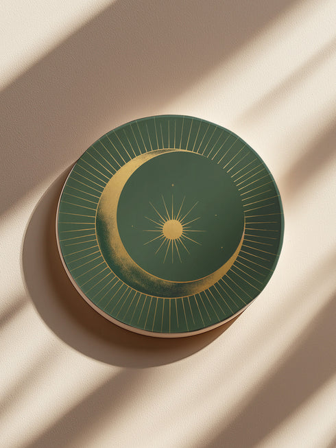 Round ceramic coaster with forest green background. Golden crescent moon embraces a sun at the center. Fine golden lines radiate outward, creating a sunburst pattern to the edge. White rim indicates cork backing. Coaster diameter is 4.04 inches.