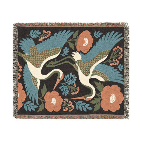 100 percent cotton woven blanket featuring a chinoiserie aesthetic. The background is a solid deep charcoal black. Scattered across the design are large flying cranes with warm cream bodies and teal blue wing feathers, alongside large rust, terracotta, and peach colored botanical flowers and olive green leaves. The textile has a visible woven texture with colorful fringed edges extending from the threads. Explicitly no purple or pink present. Curated by Grimoire and Goblet.