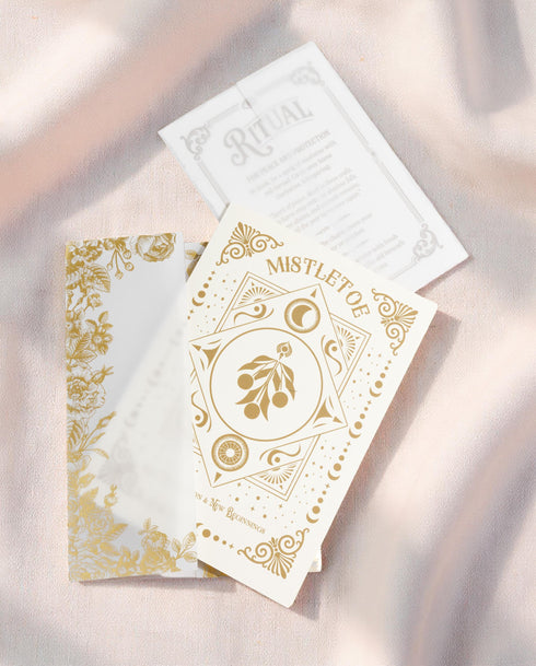 A Grimoire & Goblet luxury Mistletoe ritual card set. The 5x7 cream greeting card features gold celestial designs framing “Mistletoe” and “Love, Protection & New Beginnings.” Shown with a vellum wrap printed in gold florals, a white wax seal with mistletoe, and a premium envelope. Includes a 4x6 ritual insert on handmade deckled paper titled “Ritual for Peace and Protection.” A spiritual Yule stationery set designed as a luxurious unboxing experience.