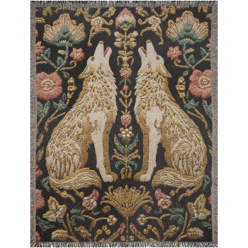 A rectangular, 100% woven cotton throw blanket with a detailed jacquard design. The throw features a symmetrical, vertical pattern of two howling wolves in profile, rendered in shades of beige, gold, and brown against a solid black background. The wolves are surrounded by intricate floral and foliage motifs. The thick, textured weave is visible. The blanket is finished on all four sides with a two-inch, multi-colored fringe that includes black, white, red, and gold threads.