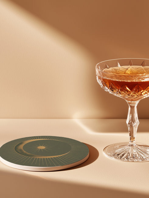 Round ceramic coaster with forest green background. Golden crescent moon embraces a sun at the center. Fine golden lines radiate outward, creating a sunburst pattern to the edge. White rim indicates cork backing. Coaster diameter is 4.04 inches.