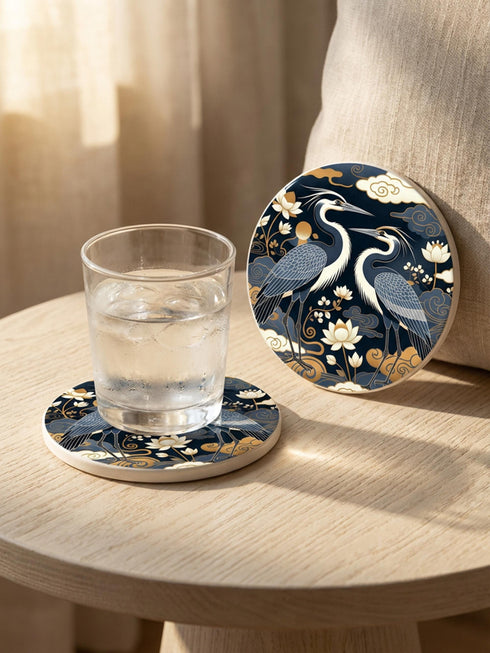 A round 4-inch drink coaster by Grimoire and Goblet. The coaster features a highly reflective glossy top surface displaying two Great Blue Herons, lotus flowers, and stylized clouds in metallic antique gold and slate blue against a solid, deep navy blue background. The edges reveal a natural brown, porous cork backing underneath. The intricate design resembles vintage Chinoiserie and botanical natural history art, perfectly suited for protecting tables from beverages.