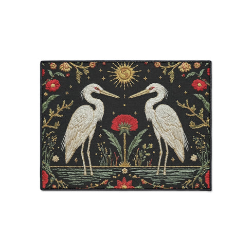 A Grimoire and Goblet rectangular doormat featuring a vintage tapestry design of two white herons facing a central red flower. Above the birds is a golden sun with a face in a celestial style. The background is a dark charcoal with a faux needlepoint texture. The scene is framed by a border of red carnations and green vines in an Arts and Crafts style. The design mimics antique embroidery but is a smooth printed surface with a black trim.