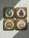 Grimoire and Goblet set of four square glossy coasters featuring high-resolution printed illustrations of raw crystals: purple amethyst, pink rose quartz, iridescent labradorite, and banded agate. Each gemstone is depicted inside a printed faux antique brass baroque frame with intricate scrollwork, set against a cream parchment background. The design is a flat 2D trompe l'œil print on hardboard, creating a realistic 3D specimen box illusion.