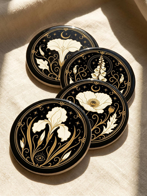 A set of four round 4-inch ceramic coasters featuring a glossy 2D flat-printed Art Deco botanical design. Each coaster displays a different cream-colored flower including a moonflower, foxglove, poppy, and iris, set against a solid black background. The floral motifs are framed by intricate gold linework, crescent moons, four-pointed stars, and mystical symbols like a candle and an all-seeing eye. The coasters have a smooth white edge and are finished with a natural brown cork backing.