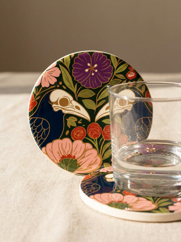 A square drink coaster with a high-gloss finish and natural cork backing. The centralized, symmetrical design features two detailed, cream-colored avian bird skulls facing inward. They are framed by lush botanical flora, including a large purple flower, blush pink blooms, rust-red roses, and green ferns, all set against a deep navy blue background with gold linework accents. Functional gothic, whimsigoth, and dark academia drinkware designed by the Grimoire and Goblet brand.