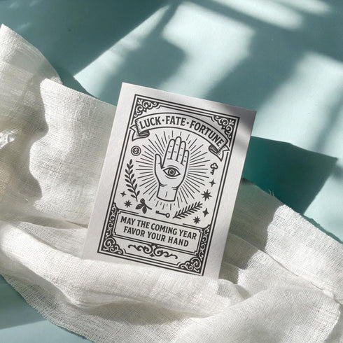 Grimoire and Goblet presents a 5x7 inch pearlescent greeting card. The design features a black linocut-style illustration of an open palm with an eye, surrounded by rays, keys, coins, and stars. The cover reads Luck Fate Fortune and May The Coming Year Favor Your Hand. The interior features a smaller hand icon and a printed blessing. The paper has a luxury silvery shimmer finish.