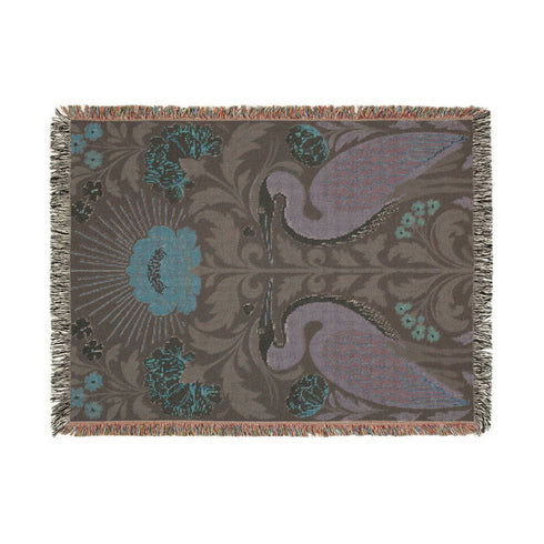 Jacquard woven cotton throw blanket, woven rather than printed, shown flat. Two mirrored great blue herons flank a central radial sunburst, surrounded by botanical blooms and scrollwork. The charcoal black ground carries antique gold ochre sunbeams, blush pink blossoms, deep forest green leaves, muted rust berry clusters, and cream toned herons. A lush multi-colored fringe of black, ochre, and sage threads edges the blanket, and the reverse shows an inverted charcoal grey version of the same design.