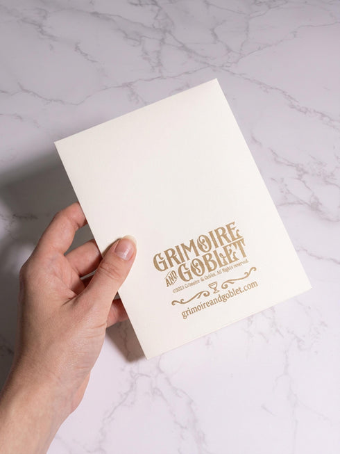 Grimoire and Goblet greeting card featuring a vintage woodcut style illustration of a radial sun with a serene human face. The sun is flanked by two Luna moths and sits above a Triple Moon symbol. A banner above reads THE MOON AGREES in gothic font. Printed in burnished gold tones on cream paper. Interior text reads Yours in this lifetime and the next. Dark Academia and Whimsigoth aesthetic stationery 5x7 card with matching envelope for gothic romance or altar decor.