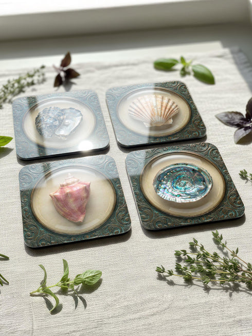 A high-fidelity 2D Trompe-l’œil print on a set of four 4x4 inch square coasters. Each coaster features a centralized sea specimen—a scallop, an oyster with a pearl, a pink conch, and an abalone—centered within an ornate, circular Victorian scrollwork frame. The design uses a faux-texture effect to mimic raised bronze and verdigris metal. The colors include aged gold, teal, and cream, creating a vintage museum specimen aesthetic