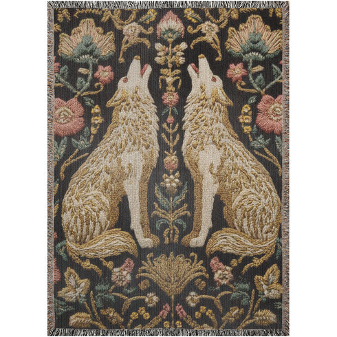 A rectangular, 100% woven cotton throw blanket with a detailed jacquard design. The throw features a symmetrical, vertical pattern of two howling wolves in profile, rendered in shades of beige, gold, and brown against a solid black background. The wolves are surrounded by intricate floral and foliage motifs. The thick, textured weave is visible. The blanket is finished on all four sides with a two-inch, multi-colored fringe that includes black, white, red, and gold threads.