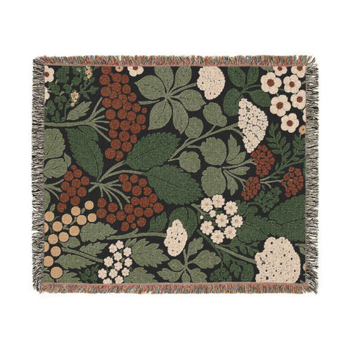 A Grimoire and Goblet luxury woven throw blanket featuring a dark botanical design. The Jacquard cotton tapestry showcases cream elderflower clusters and rust-colored berries against a deep black background. Sage green leaves interweave throughout the floral pattern. The blanket is finished with multicolored fringe edges. A moody cottagecore aesthetic piece ideal for dark academia decor.