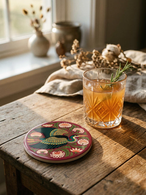 A round, 4-inch ceramic drink coaster featuring a glossy, flat-printed Art Nouveau design. The central motif is a flying heron with teal plumage and scalloped golden wing feathers, surrounded by stylized winding olive green foliage, peach chrysanthemums, and botanical blooms. The artwork is set against a solid, deep blood-red background. The coaster has a smooth white edge and a natural brown cork backing. Available as a single coaster or in matching sets of four or six.