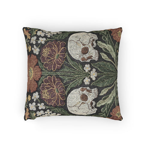 A Grimoire and Goblet woven tapestry pillow featuring a symmetrical dark botanical design. Two beige human skulls face each other in profile against a dark charcoal background. They are surrounded by deep burgundy dahlia flowers, sage green leaves, and small white floral clusters in an Arts and Crafts style. The texture is a visible fabric weave. The design is double-sided, square, and filled.